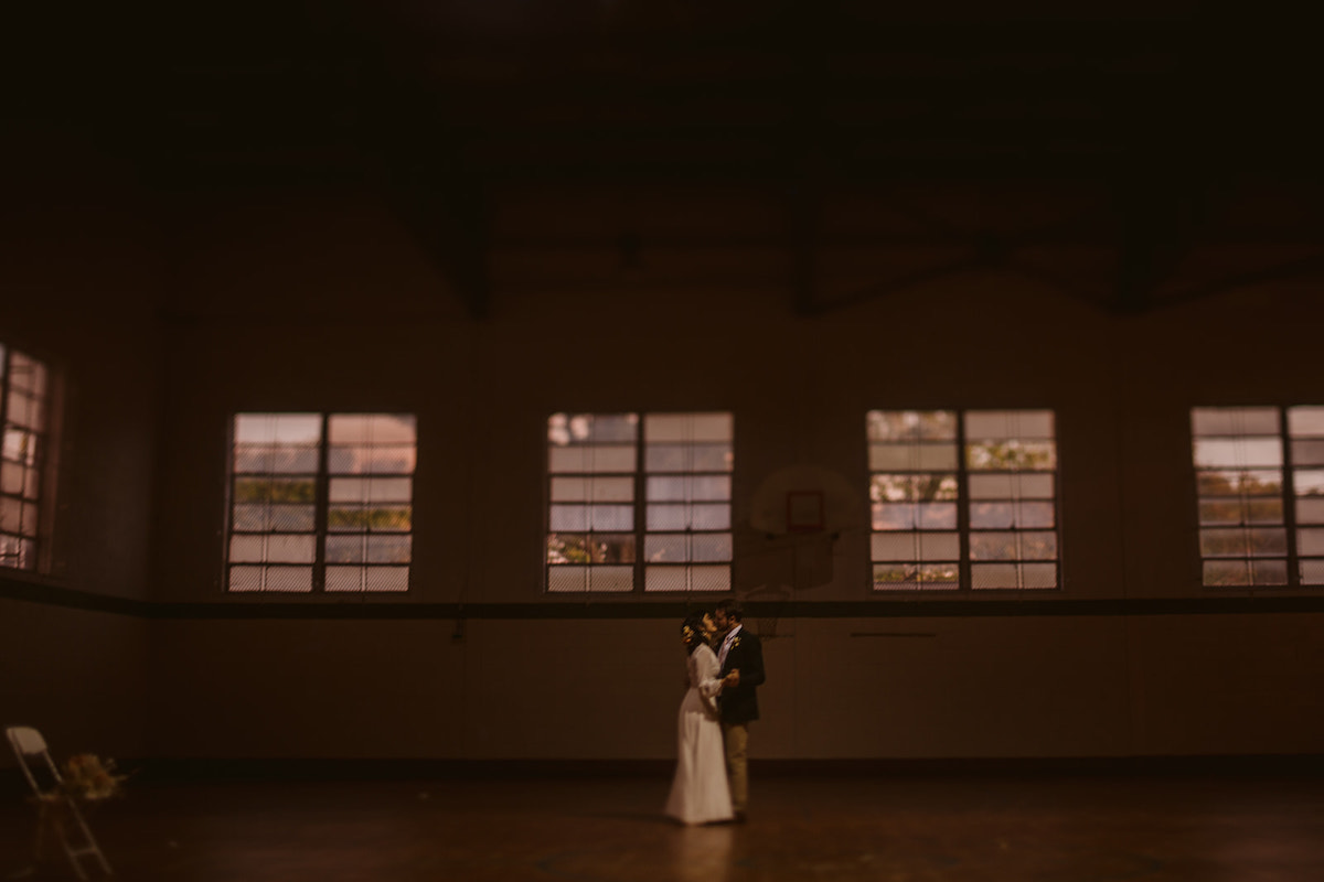 bride and groom dance together under a basketball hoop in a shadowy gymnasium