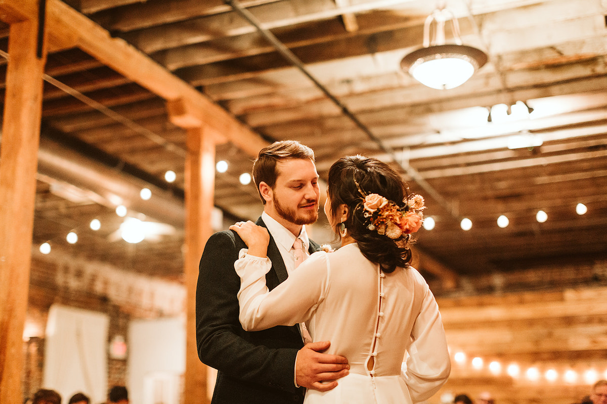 Groom looks at bride share their first dance under exposed rafters and beams during their reception at The Old Woolen Mill