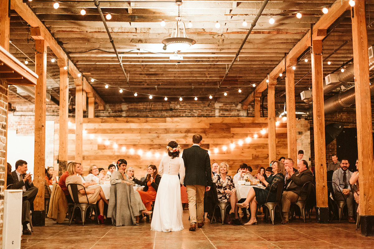 Bride and groom stand before their wedding guests under string patio lights across exposed rafters of a restored warehouse