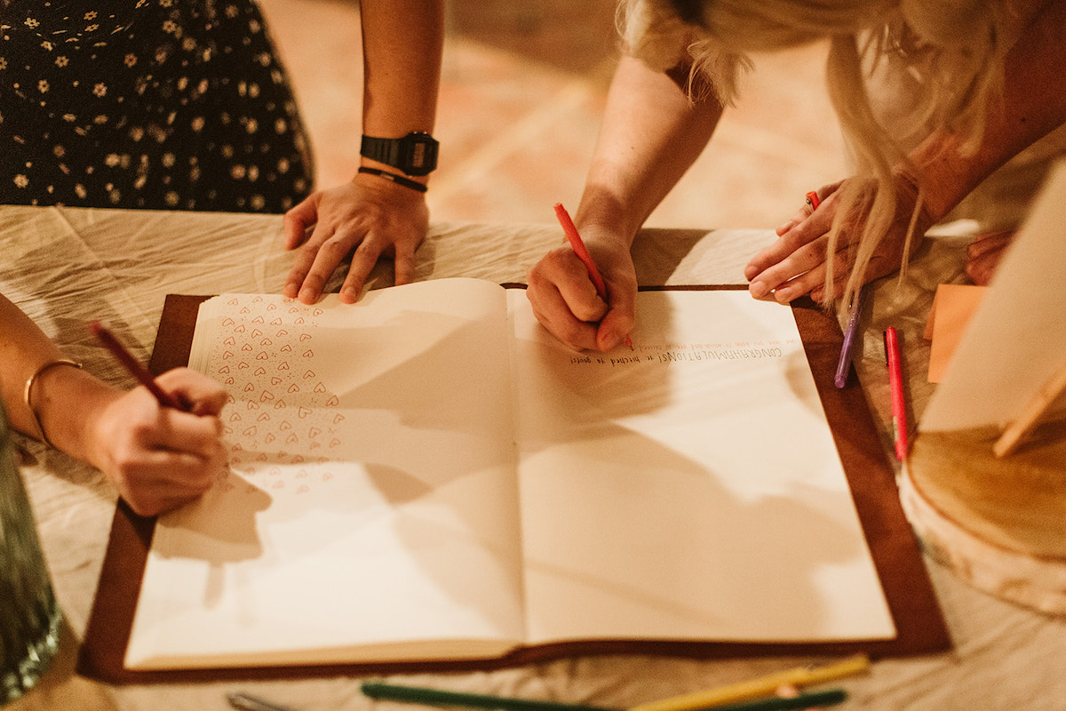 Two women sign the large blank pages of a guest book, one fills the page with hearts