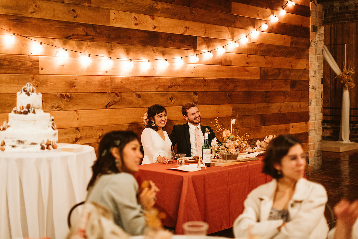 Bride and groom sit at a table with amber-colored tablecloth in front of a shiplap wall strung with patio string lights