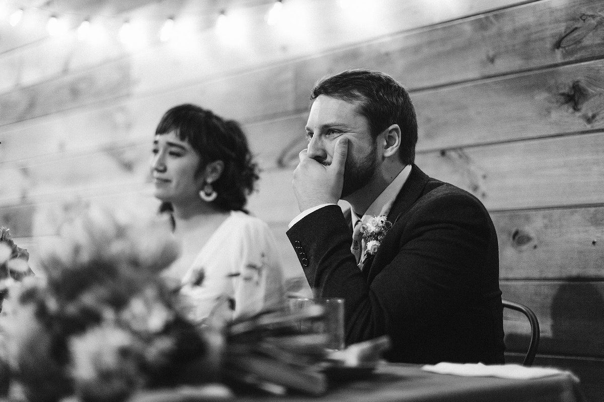 Bride and groom sit in front of shiplap wall at The Old Woolen Mill. He holds his mouth emotionally.