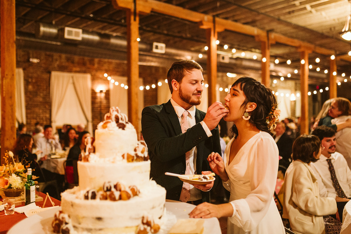 Groom hand-feeds bride cake next to their 3-tier cake topped with cream puffs