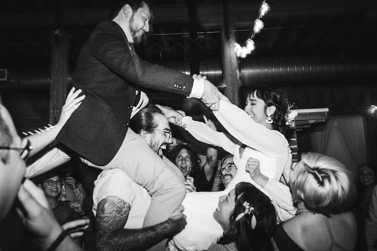 Bride and groom hold onto each other's hands as they sit on top of their friends' shoulders