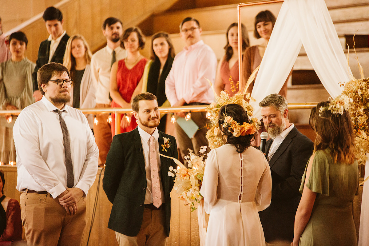 Groom smiles at bride, his best man behind him and several wedding attendants watch from gym bleachers