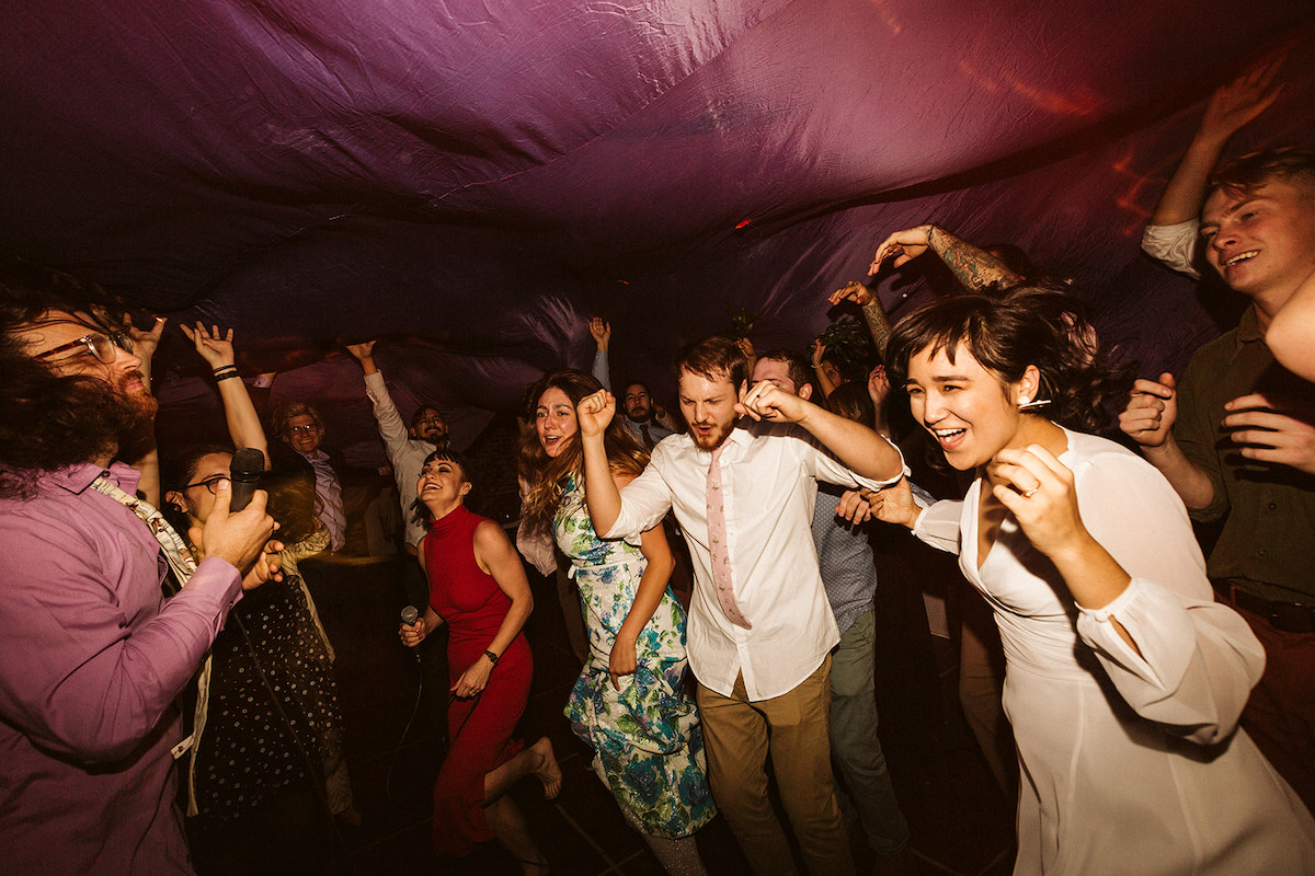 Bride and groom dance and jump with friends under a large purple parachute