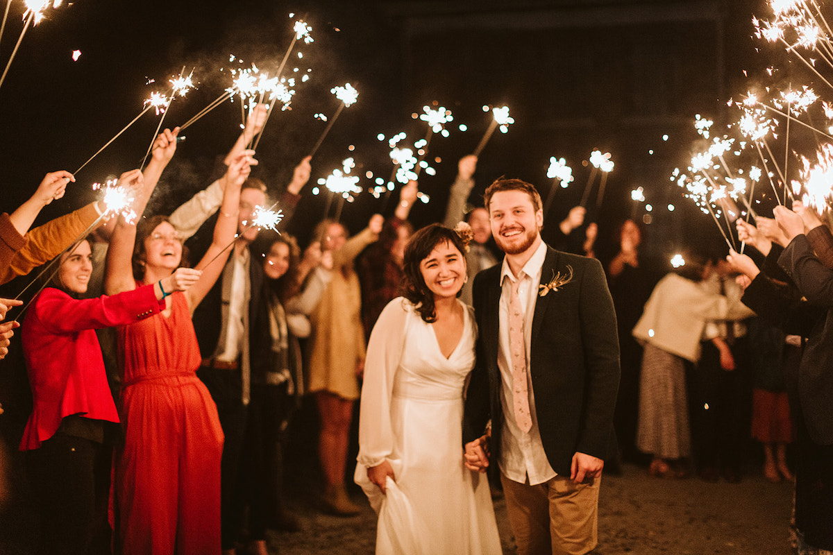 Bride and groom walk between rows of their friends extending sparklers into the night sky