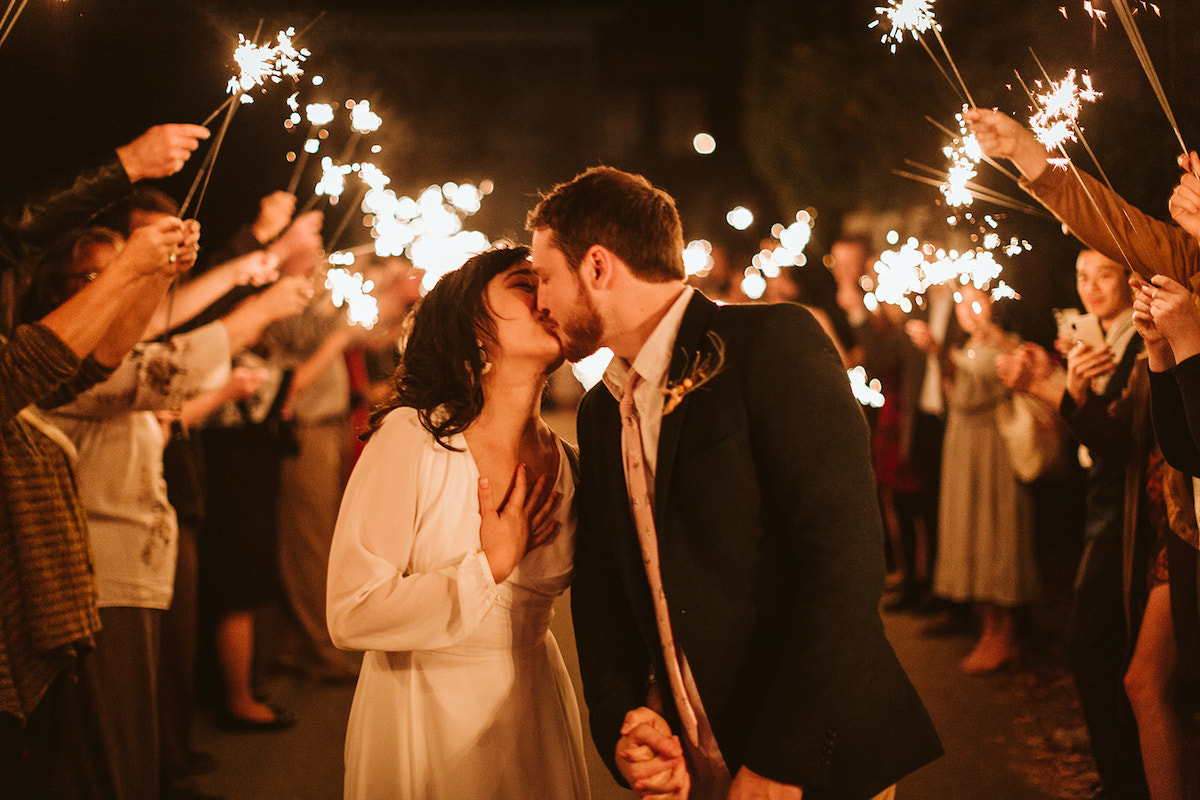 Bride and groom hold hands and kiss under sparklers, she holds a hand over her heart