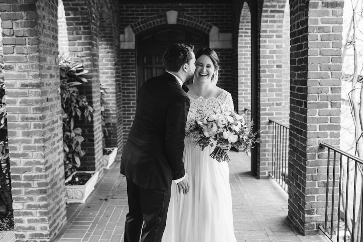 Groom kisses bride's cheek under covered walkway between brick archways