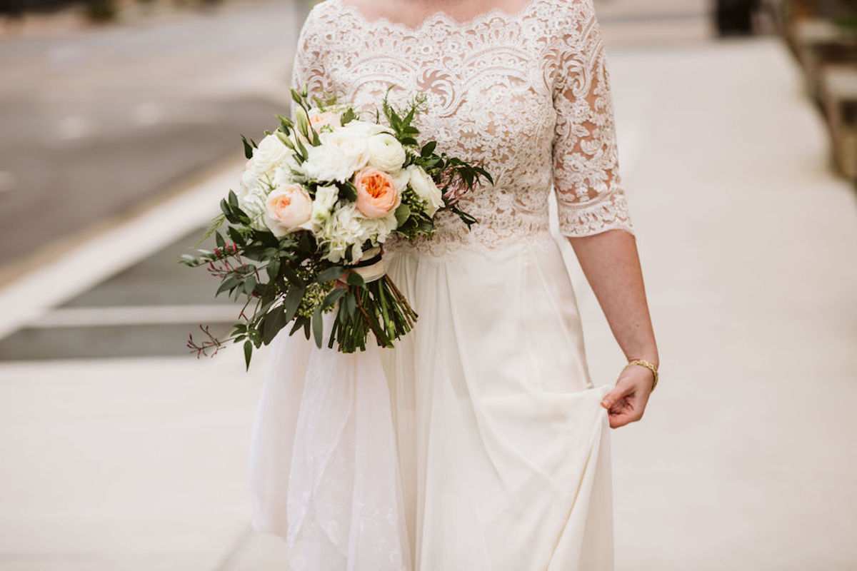 Bride wears high-collared, three-quarter-length lace wedding dress holds bouquet of white and peach flowers