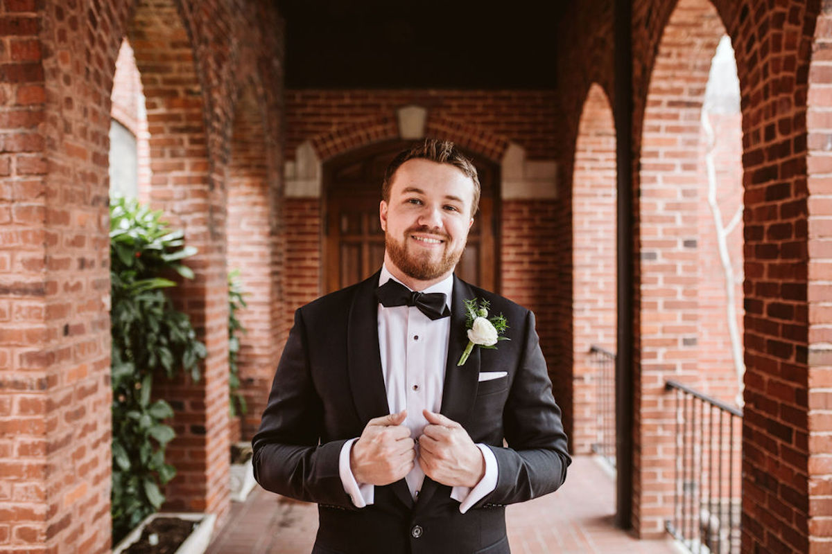 Groom holds lapels of his tuxedo with a white boutonniere pinned on his chest