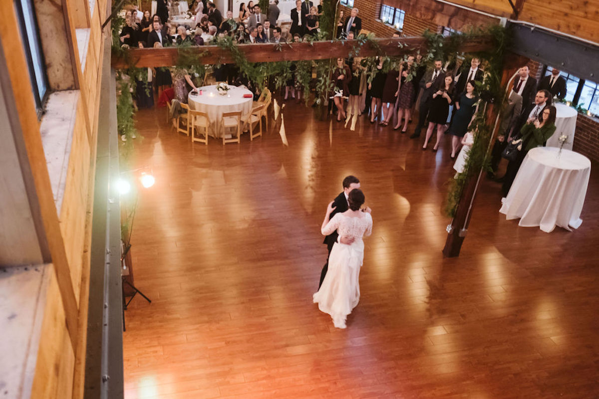 Bride and groom dance alone on wooden dance floor under exposed beams of The Turnbull Building while guests watch
