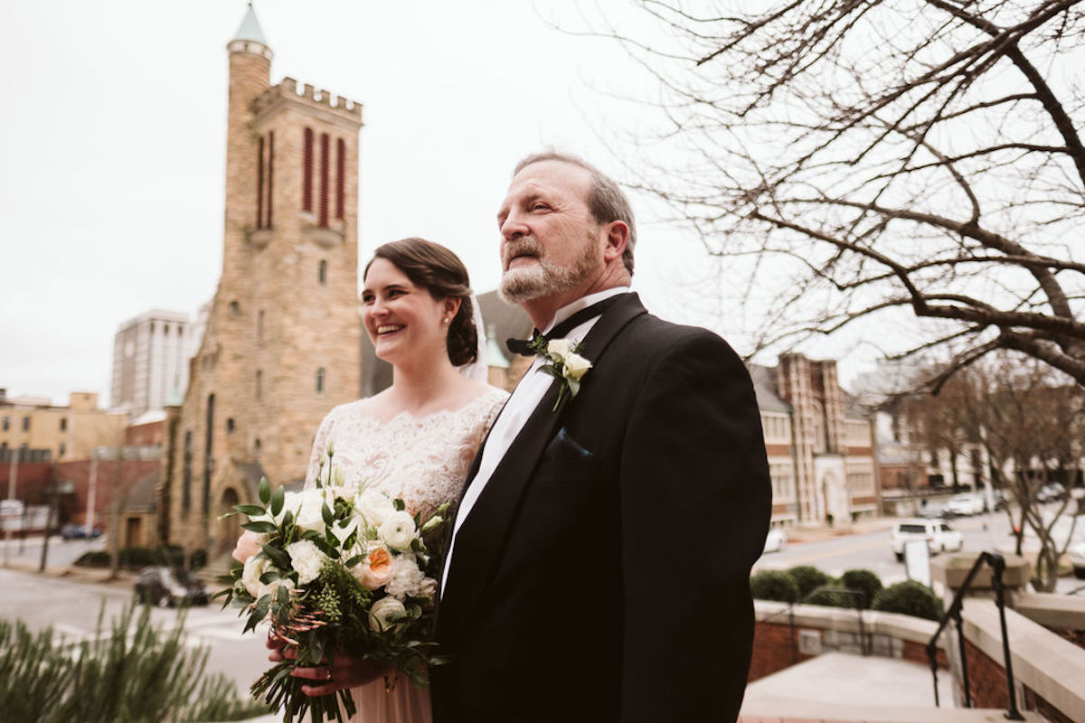 Bride and her father stand under gray winter sky in front of antique brick church