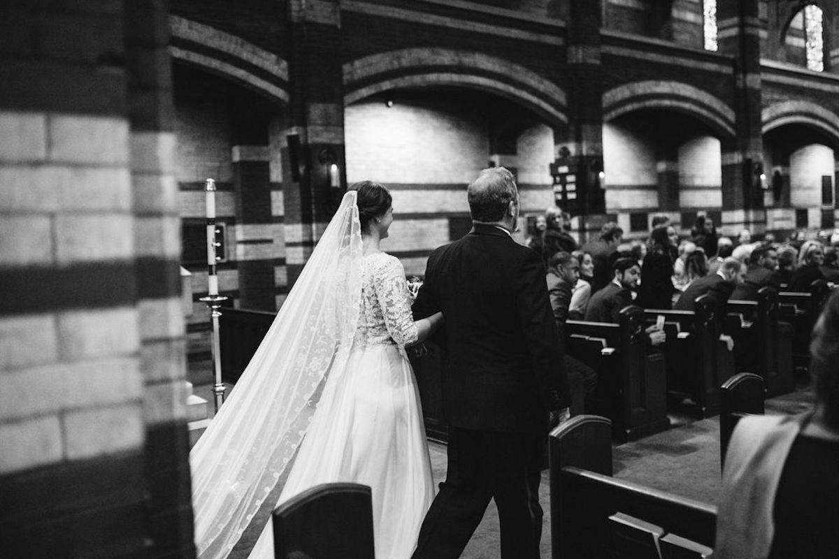 Bride and father walk central aisle as guests begin to stand from wooden pews for processional
