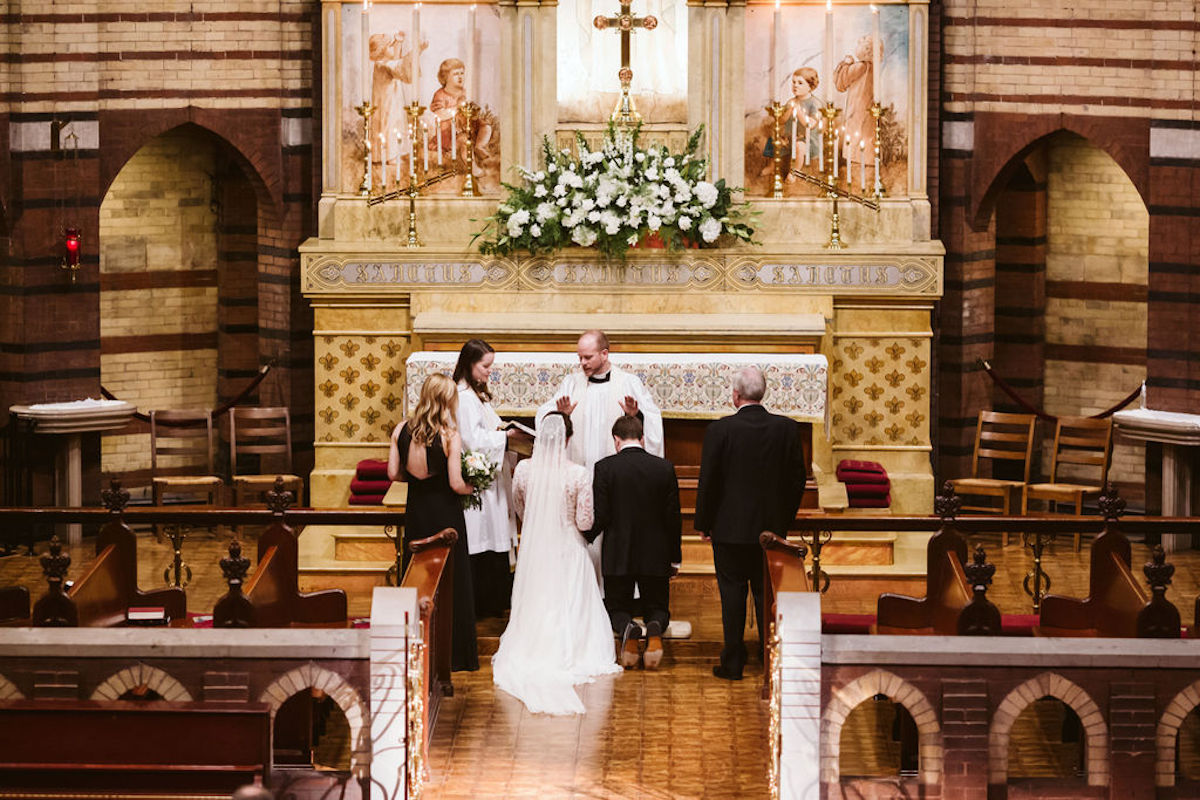 Bride and groom kneel at altar while priest reads Scripture during their wedding ceremony
