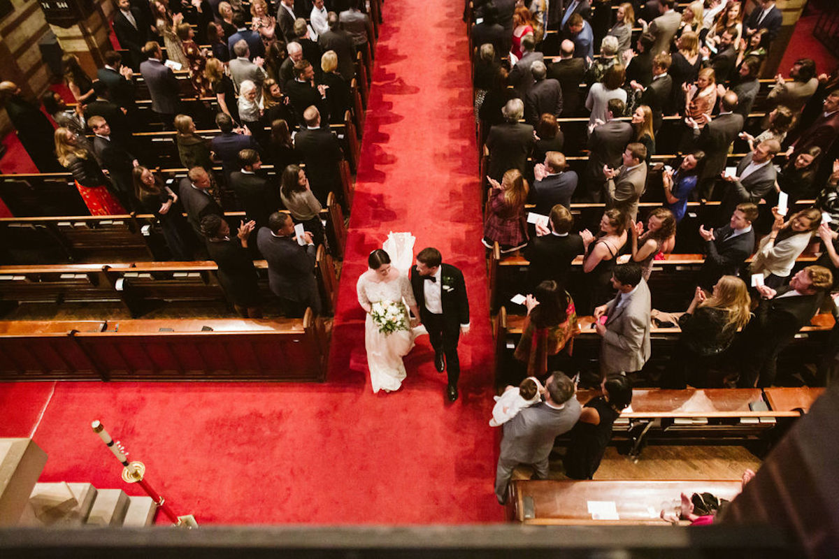 Bride and groom hold hands and walk the red-carpeted aisle while guests stand and clap