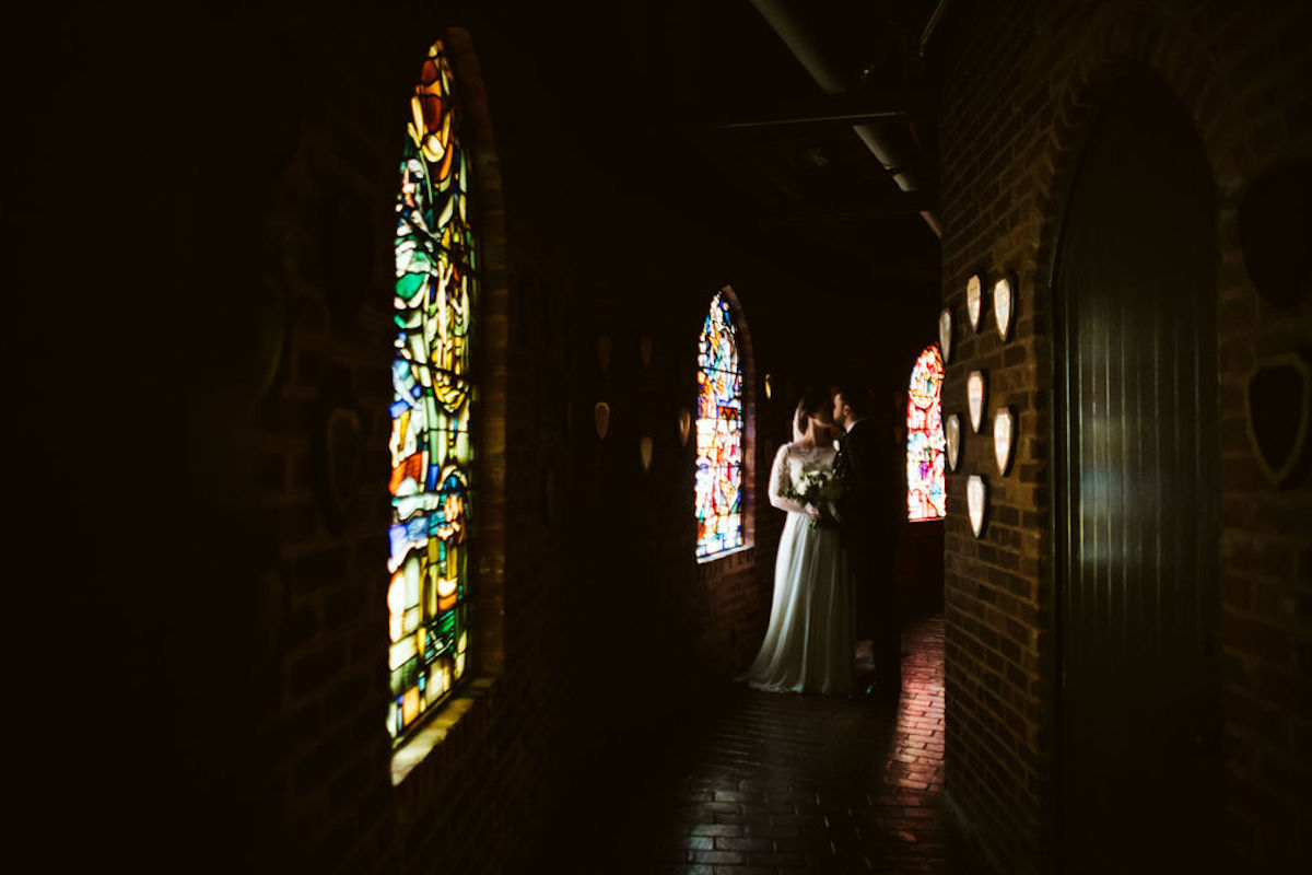 Bride and groom kiss in shadowy hallway between stained glass windows