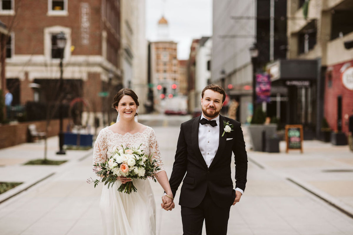 Bride and groom hold hands in Chattanooga's West Village. She holds a bouquet of white and peach flowers