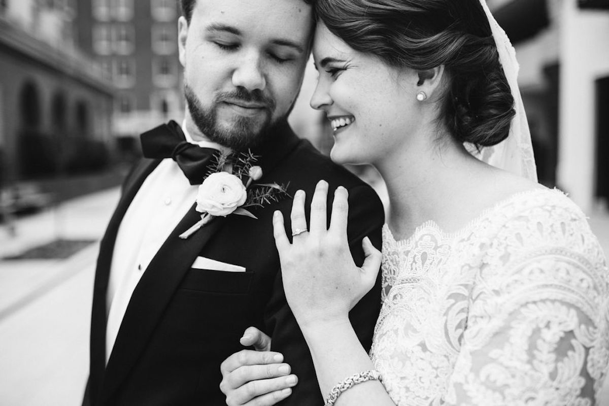 Bride and groom cuddle, her hand under his arm and the other on his shoulder.