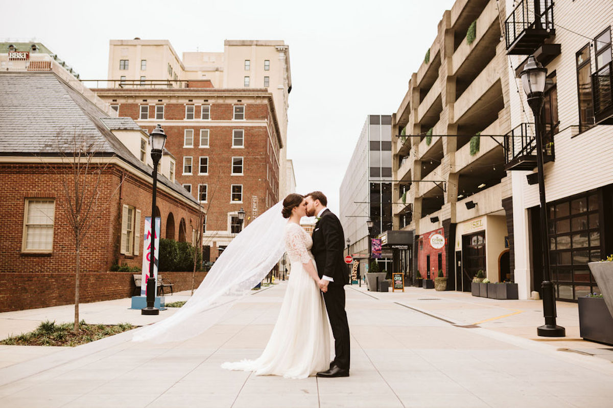 Bride and groom kiss in the middle of the road between downtown buildings. Her veil blows in the wind.