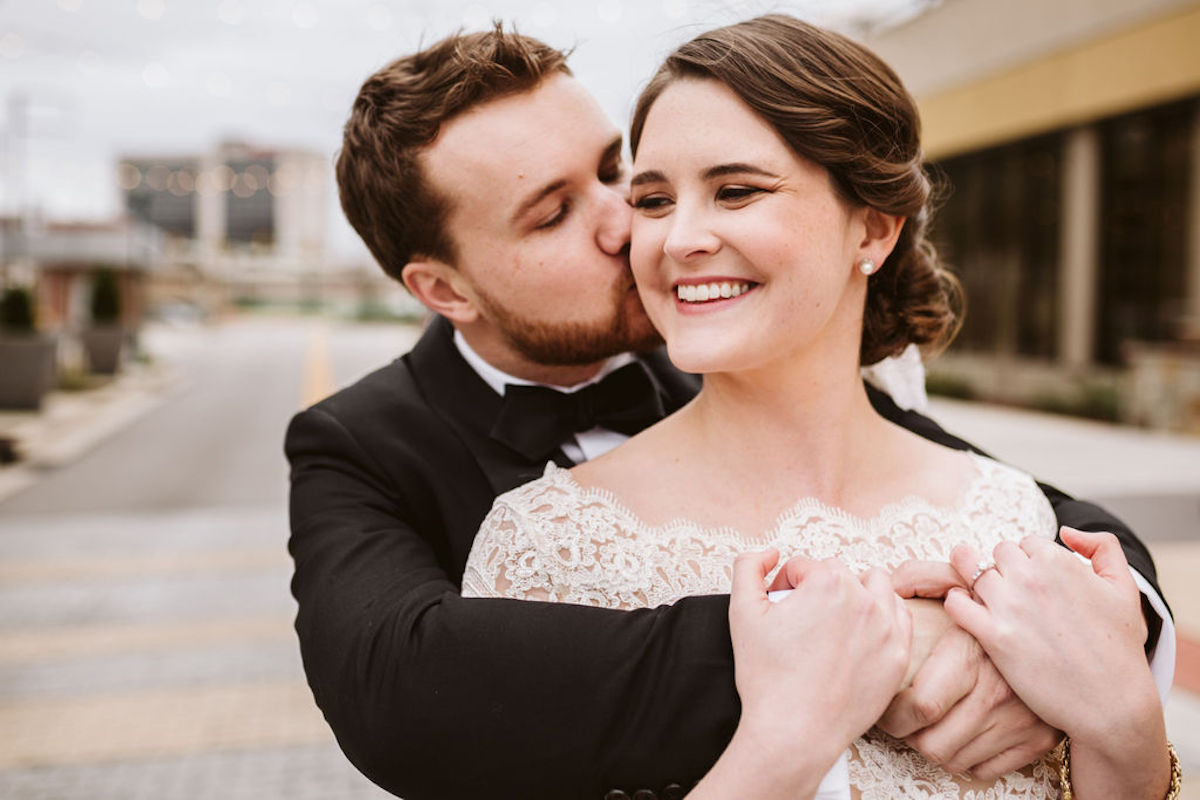 Groom cuddles bride over her shoulders while he kisses her cheek