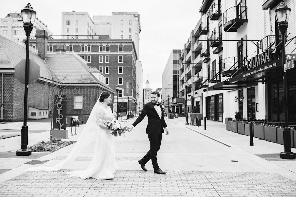 Bride and groom hold hands and cross the street in West Village Chattanooga. She holds a bouquet of white and peach flowers