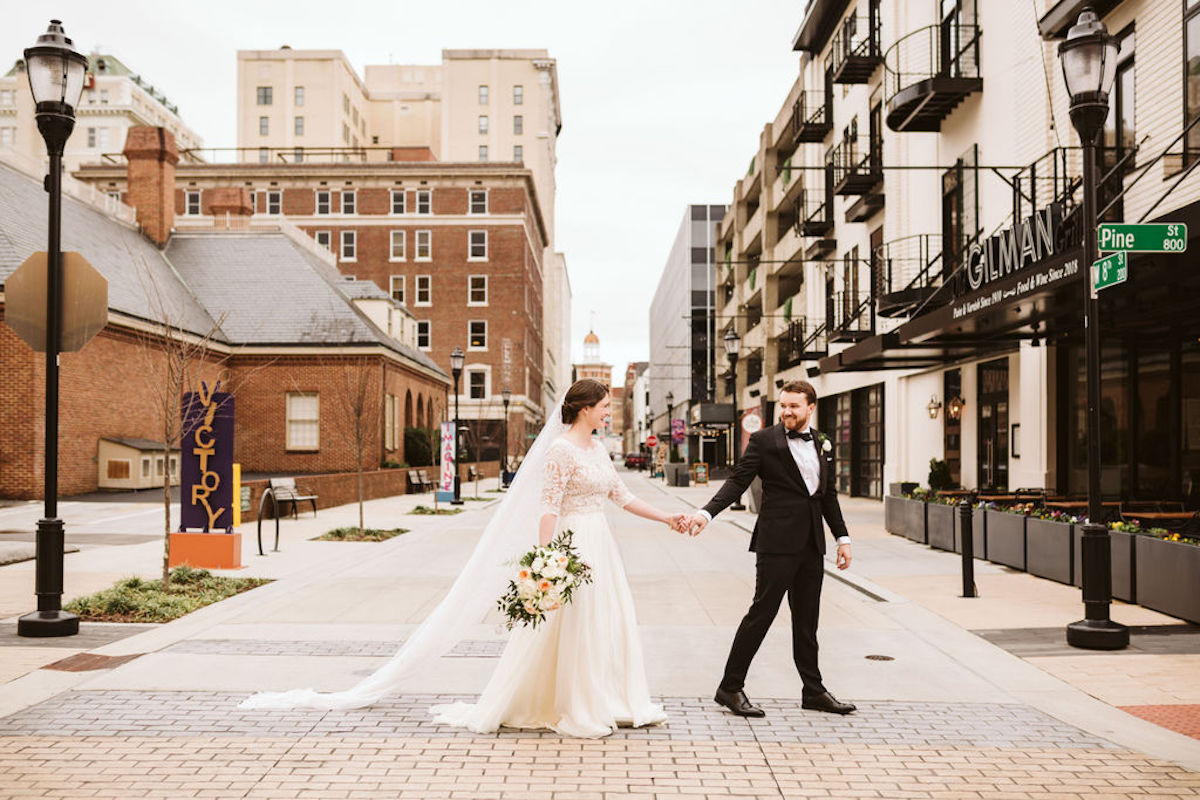 Bride and groom hold hands and cross the street in West Village Chattanooga. She holds a bouquet of white and peach flowers