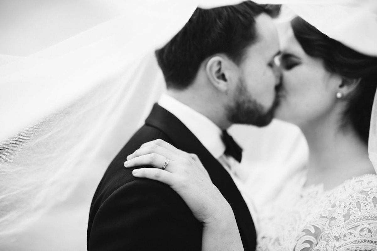 Bride and groom kiss under her veil, her ring hand on his shoulder.