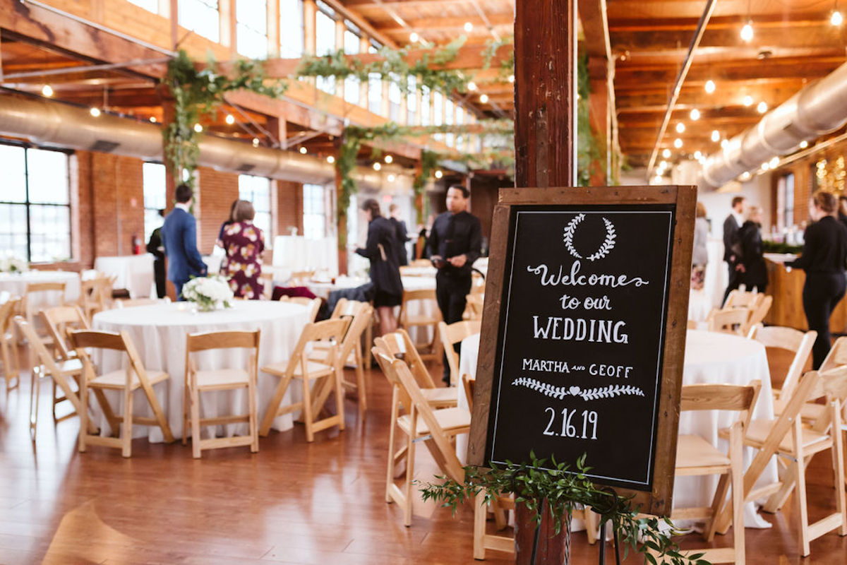 Chalkboard in front of round tables with white tablecloths under large windows and exposed beams at The Turnbull Building