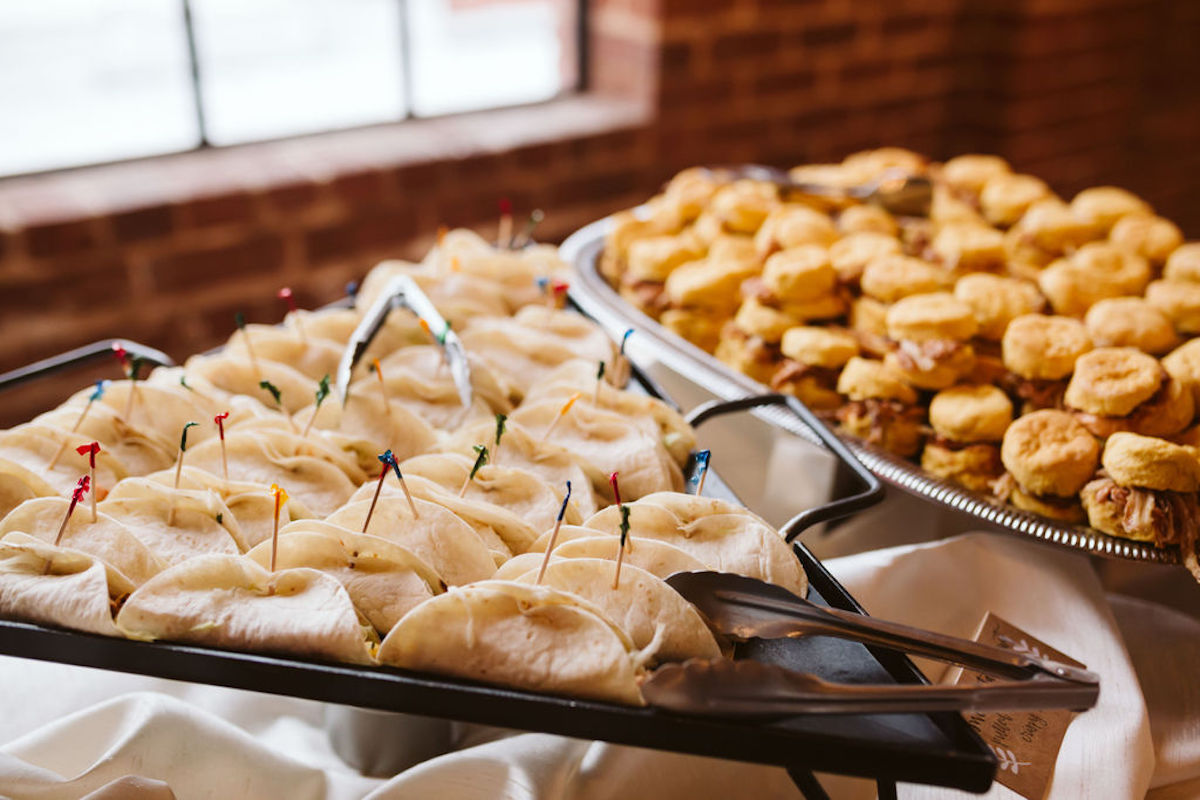 Serving tongs on a platter with soft shell tacos next to a platter of pulled pork sliders