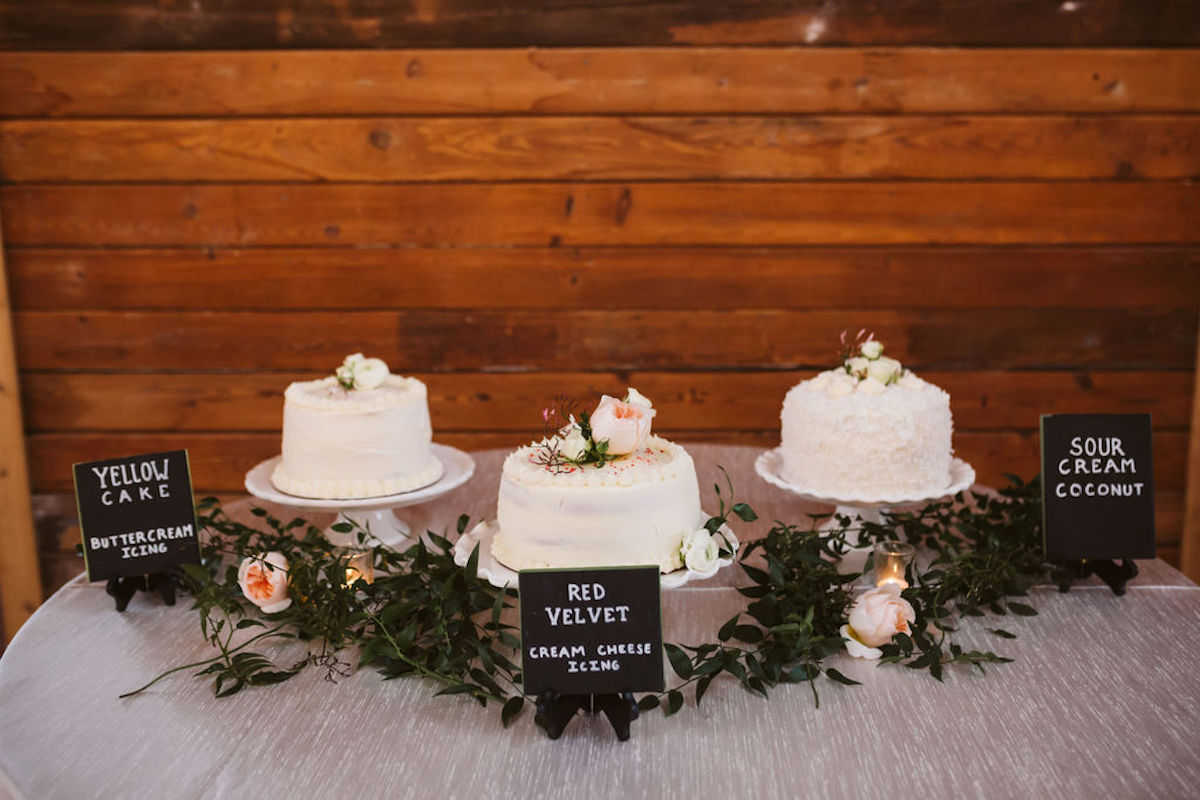 Three white frosted layer cakes surrounded by greenery and tabletop signs labeling yellow, red velvet, and sour cream cakes