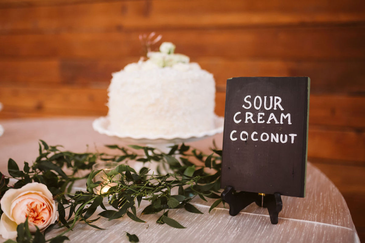 Tabletop sign printed with "Sour cream coconut" in front of greenery and a white cake topped with flowers