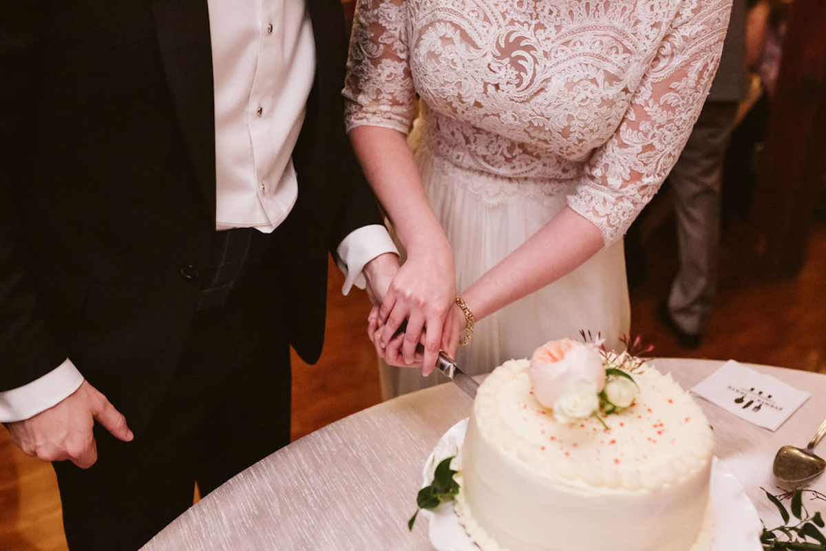Bride's hands surround groom's as they cut into their white frosted cake with pink peony on top