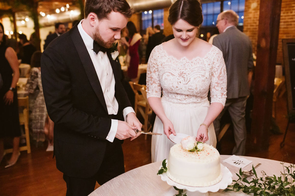 Bride holds a small plate while groom holds knife in front of white frosted cake