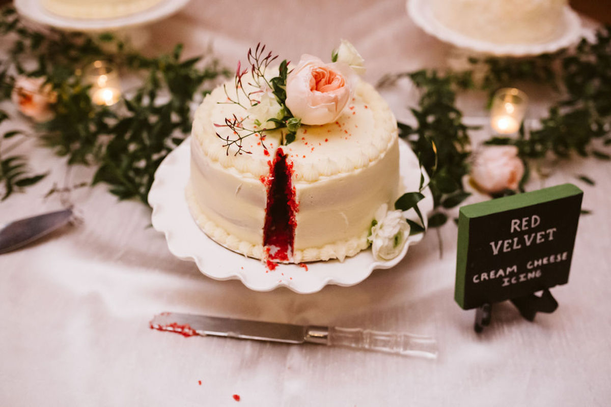 Red velvet cake with thick white frosting with small piece cut away is topped with light pink peony