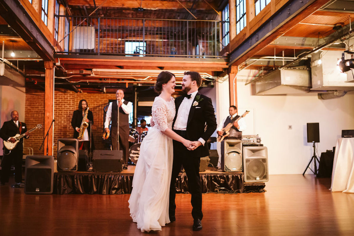 Bride and groom dance in front of live band under exposed beams and large windows at their wedding at The Turnbull Building