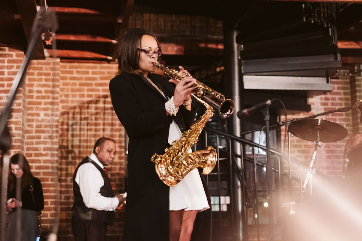 Woman plays soprano sax into microphone while tenor sax suspends around her neck