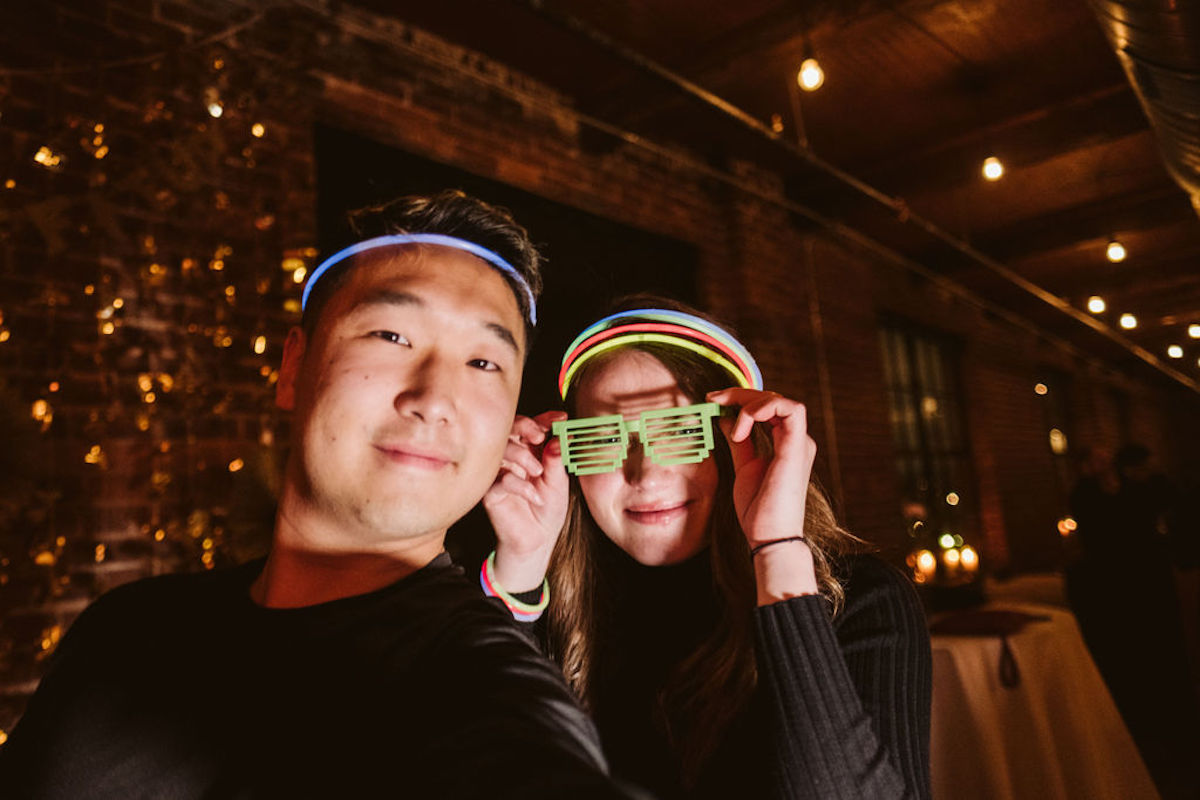 Man and woman wearing glow sticks as halos. She holds neon green glasses in front of her eyes