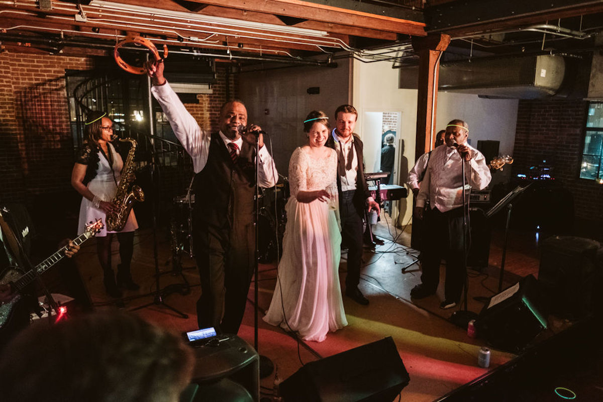 Bride and groom onstage with wedding band. The singer holds a microphone in one hand and tambourine overhead with the other
