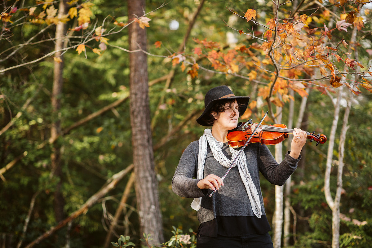 Woman in gray and black plays a violin under autumn colored trees