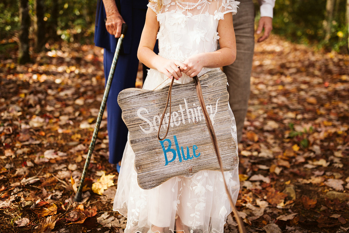 Small girl in white dress carries a wooden sign with "something blue" painted on it