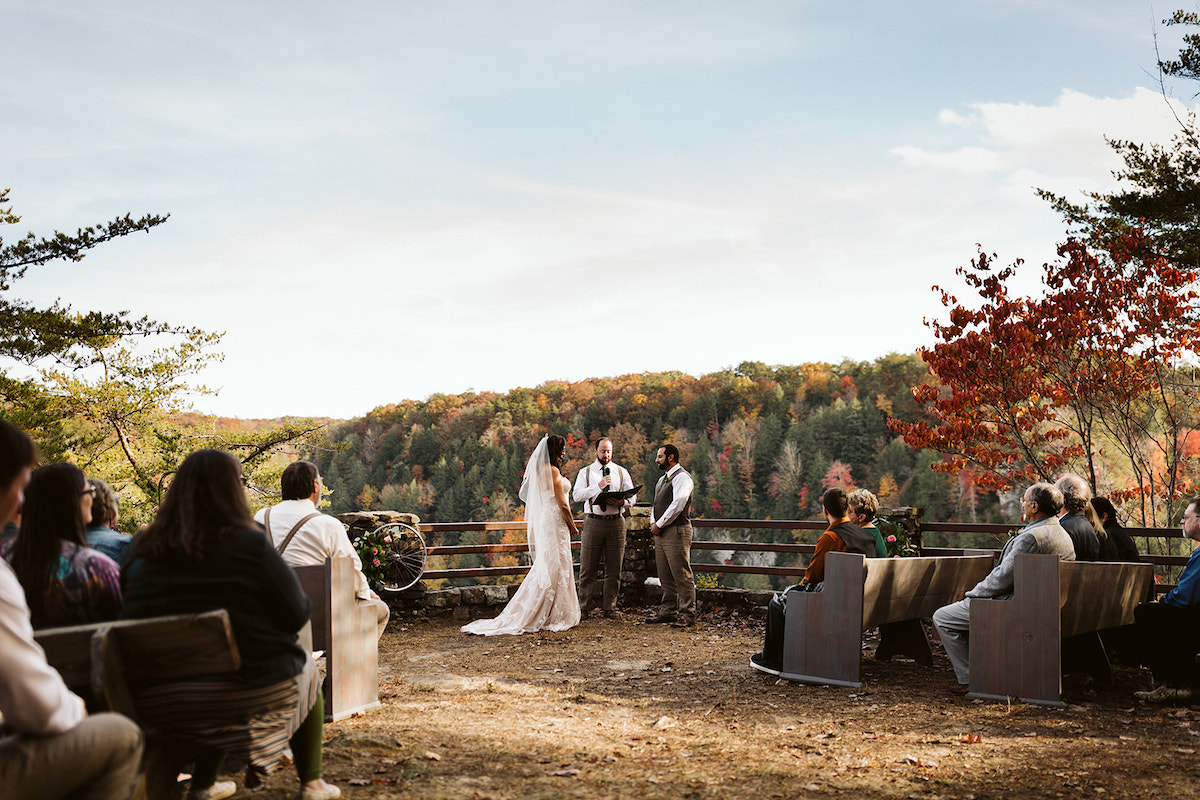 Groom and bride stand with officiant near a fence and stone pillar at an overlook at Fall Creek Falls State Park in eastern Tennessee