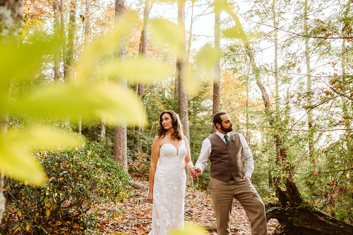 bride and groom hold hands and stand on a leaf-covered trail under tall trees with colorful leaves
