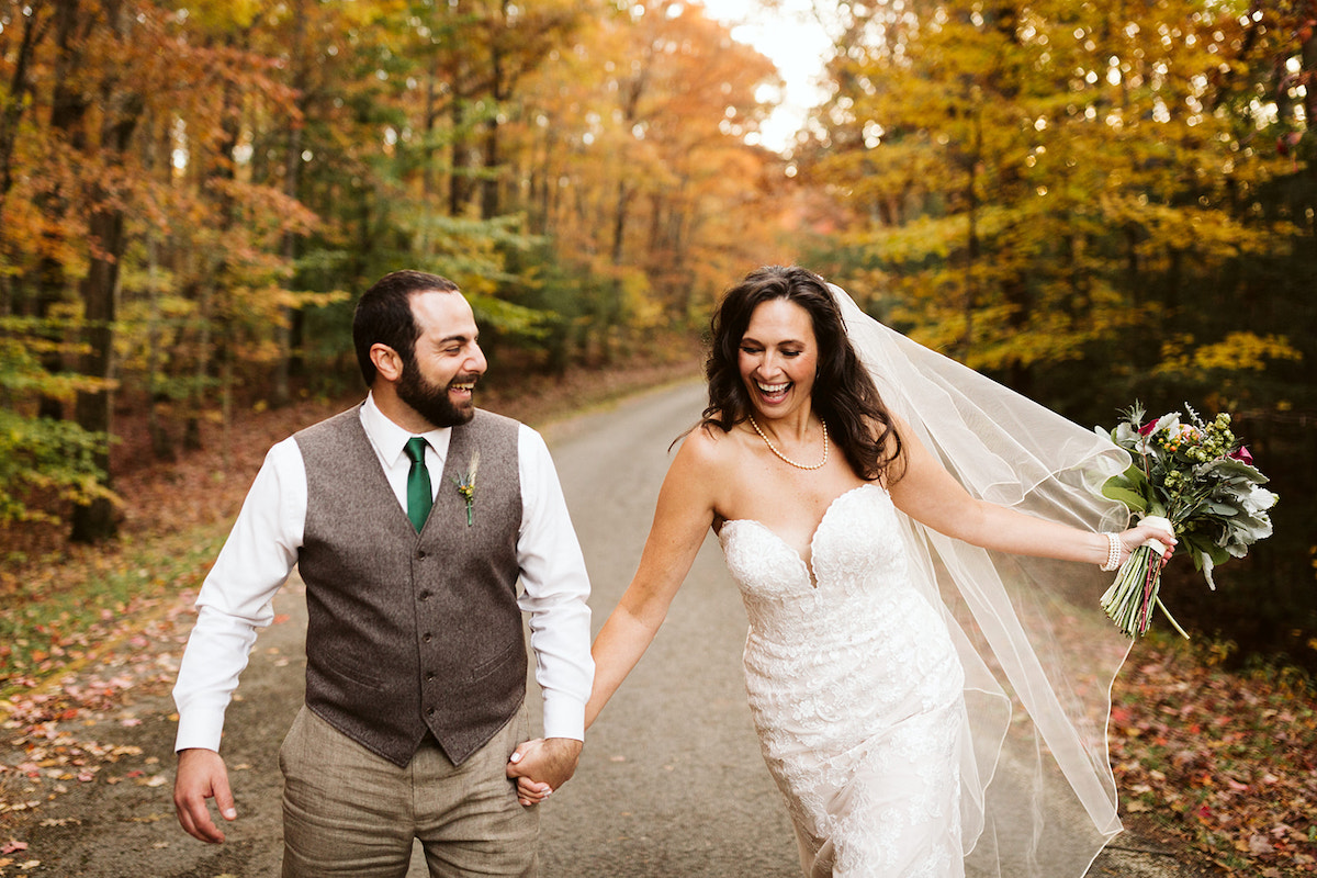 bride and groom hold hands and laugh and walk along a paved road lined with colorful trees