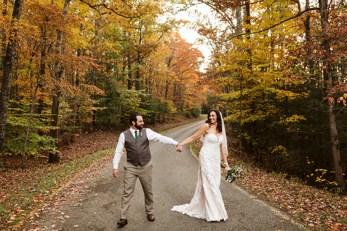 bride and groom hold hands and laugh and walk along a paved road lined with colorful trees