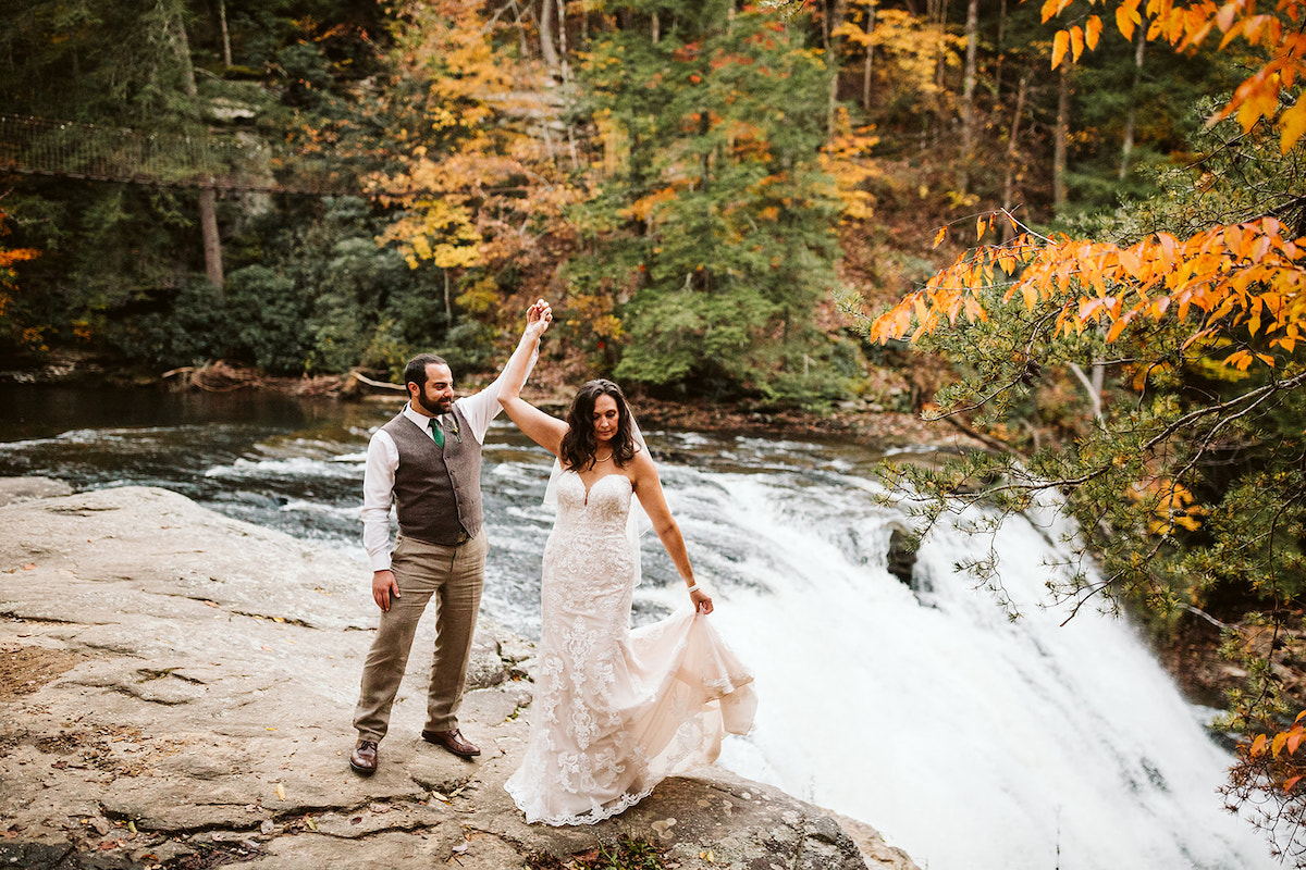 Groom twirls bride beside a rushing waterfall at autumn Fall Creek Falls State Park wedding