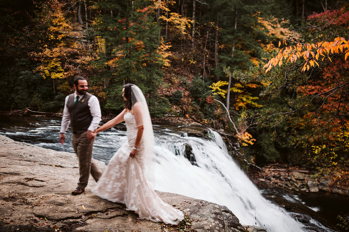 Groom and bride stand beside a rushing waterfall at autumn wedding at Fall Creek Falls State Park in Tennessee