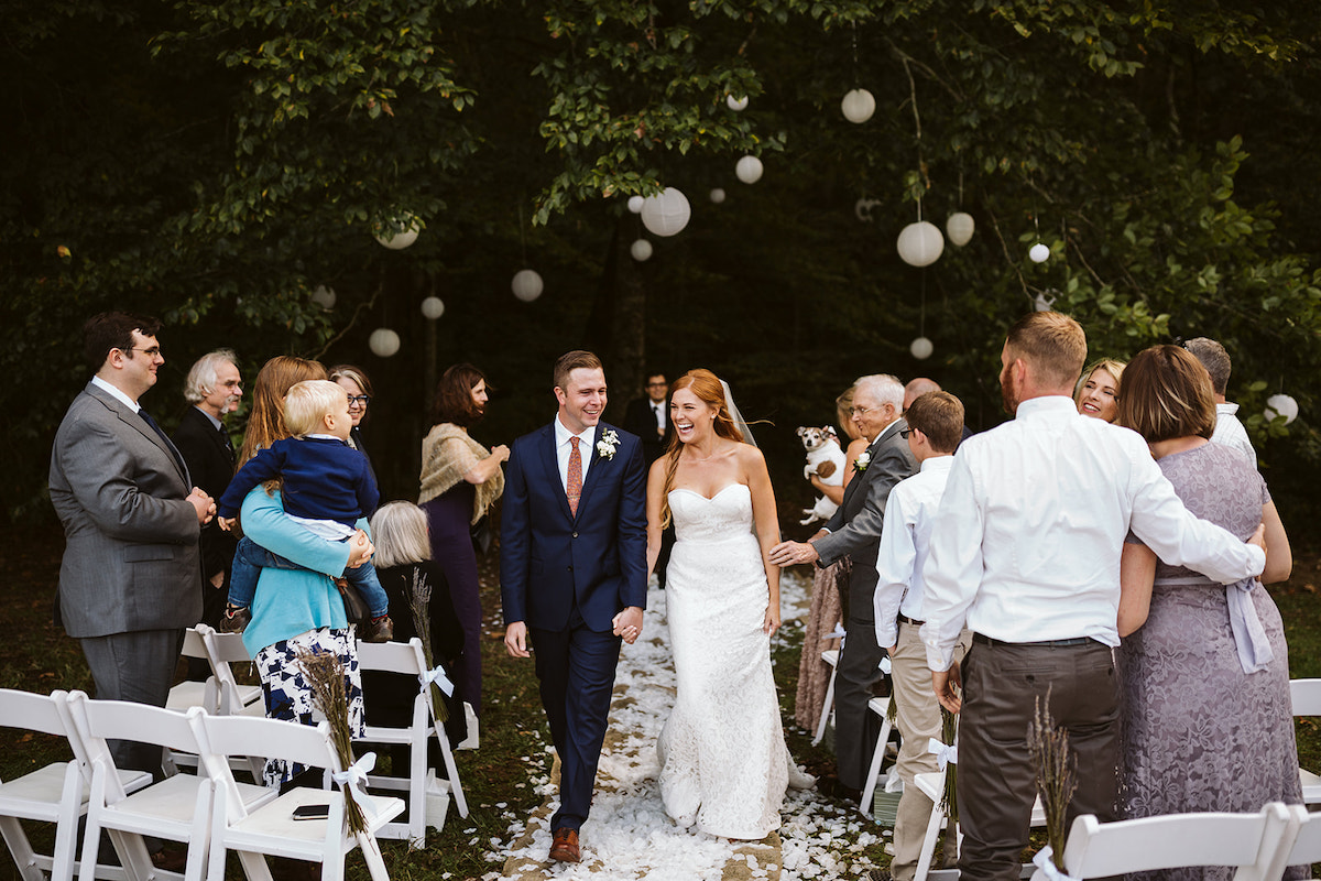 bride and groom walk between white folding chairs, holding hands. A large tree strung with white paper lanterns is behind them.