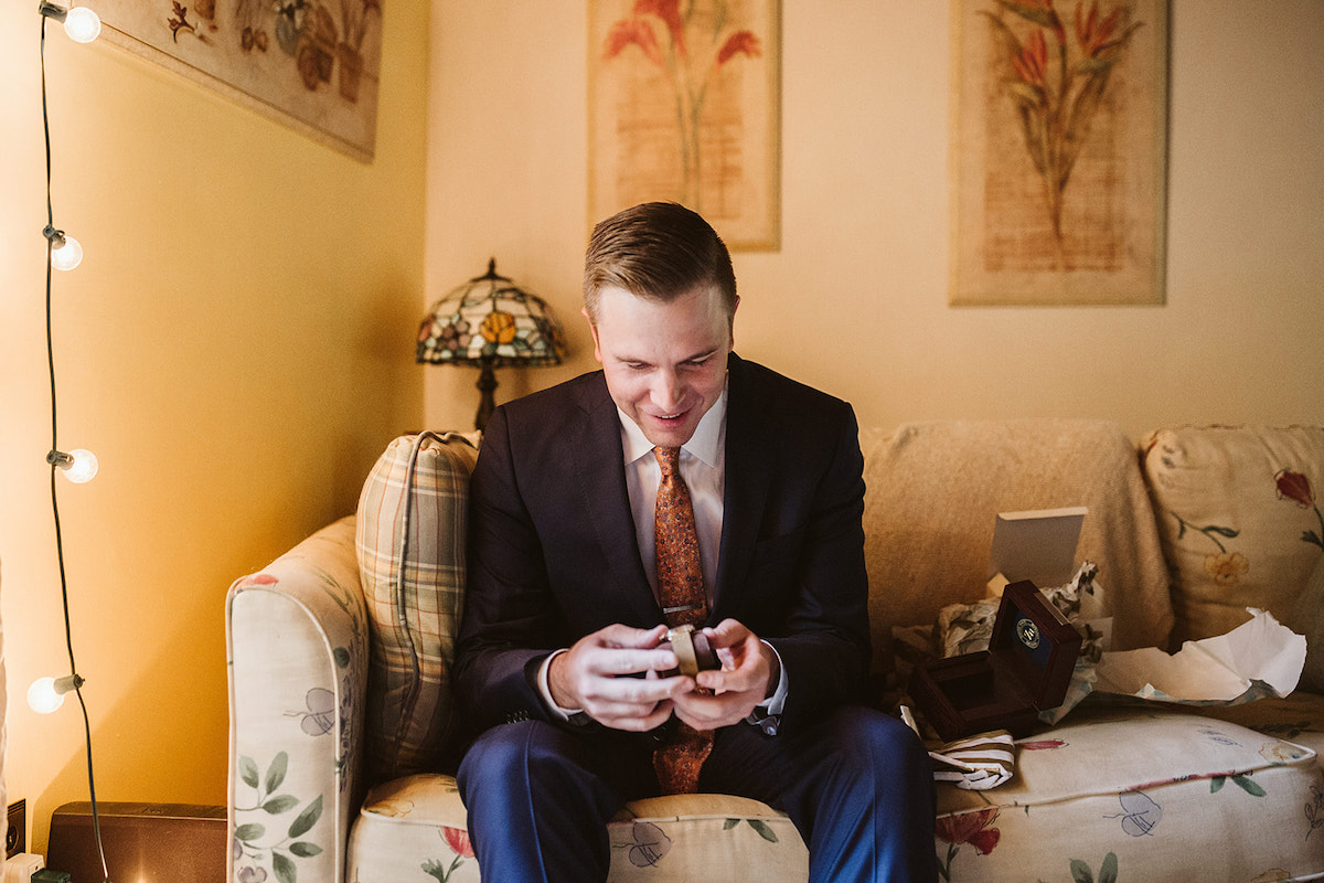 groom sits on floral couch and opens a watch gift box