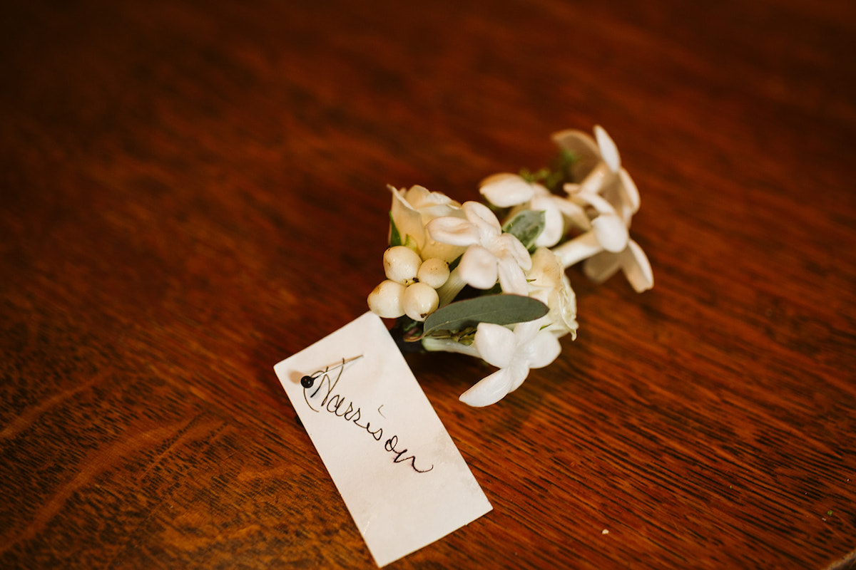 boutonniere with small, delicate white flowers. white tag with "Harrison" handwritten on it is pinned to the boutonniere